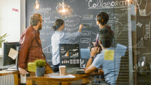 Diverse Team Of Young Developers Draw Work Plan On A Blackboard Wall, Have Heated Discussion. Creative Office Space With Stylishly Dressed People.