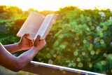 Fototapeta  - Close-up of man's hands holding a book by the window for reading against sunset with beautiful nature background. warm tone and soft focus.
