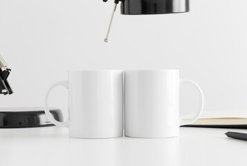 Two white mugs mockup with a lamp and a notebook on a white table.