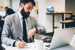 © BullRun - Handsome male lawyer looking at wristwatch while writing down information in notepad sitting at digital computer.Bearded economist in formal wear checking time on smartwatch to finish work on time