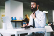 © BullRun - Confident bearded office manager in stylish black tie watching database on modern computer device and making calculations of financial reports sitting in office building near publicity area