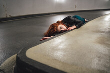 Red Haired Female Model Laying On Floor Inside Parking House Along Curb. Young Woman With Open Eyes Acting Dead Victim Of Accident.