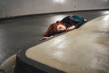 Red haired female model laying on floor inside parking house along curb. Young woman with open eyes acting dead victim of accident.