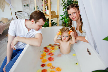 Cute little girl sitting in a bath with milk and fruit. A young family mother, father and daughter spend time together. The concept of a family holiday. Bathing a child.