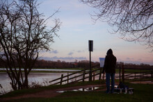 A Painter Is Standing In Front Of Easel And  Empty Canvas Praparing To Paint A Landscape Of The Scenic Port Meadow By River Thames, At Sunset. Artist Wears Winter Clothes.