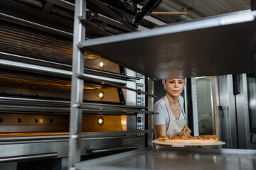 Wall Mural - Young caucasian woman baker is holding a wood peel with fresh pizza near an oven at a baking manufacture factory and putting it on a metal shelves rack.