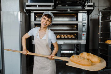 Wall Mural - Young caucasian woman baker is holding a wood peel with fresh bread near an oven at baking manufacture factory.