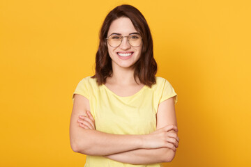 Young attractive brunette girl wearing casual clothing, having beautiful dark hair, looking at camera with toothy smile, lady standing against yellow background with folded hands.