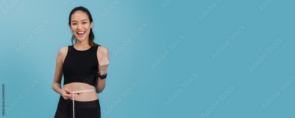 Portrait of confident beautiful asian fitness woman standing and use tape measure to measure waist  after exercise with smiling face on blue colour background.Concept of slim and healthy happy girl. - obrazy, fototapety, plakaty 