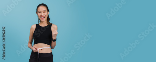 Photographie Portrait of confident beautiful asian fitness woman standing and use tape measure to measure waist after exercise with smiling face on blue colour background