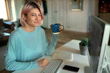 People, occupation, electronic gadgets and freelance concept. Cute young female graphic designer working on visual content for website, using desktop computer, having tea, smiling at camera