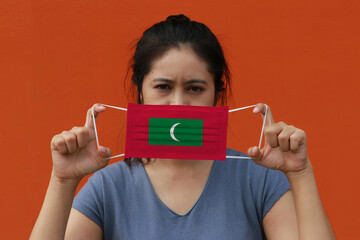 A woman with Maldives flag on hygienic mask in her hand and lifted up the front face on orange color background. Tiny Particle or virus corona or Covid 19 protection.