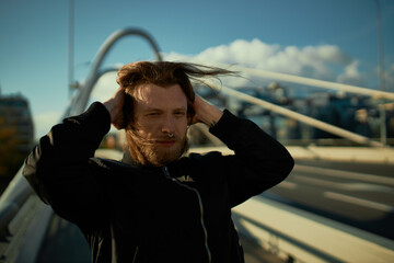 Wall Mural - Outdoor image of fashonable young male hipster witb thick beard and long hair posing on modern bridge, covering ears with hands because of strong wind. Stylish guy with nose ring walking in cityscape