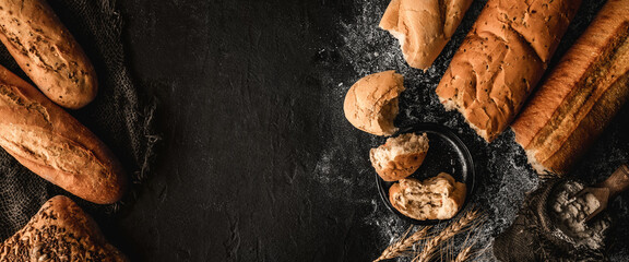 Wall Mural - Freshly bread baguettes and buns on black slate background with wheat spikelets, sackcloth. Pastries and bakery, top view, wide composition