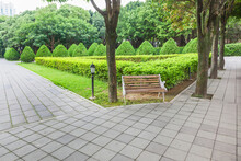 View Of Green Garden With A Small Square And Wooden Bench For Resing In The Corner Of The City Park
