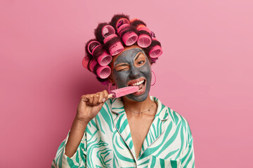 Wall Mural - Portrait of cheerful woman with clay nourishing mask on face, bites delicious ice cream with appetite enjoys her favorite frozen dessert uses hair curlers for making hairstyle on party dressed in robe