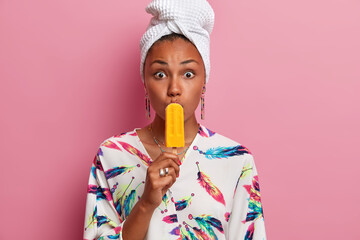 Wall Mural - Cheerful summer mood and dessert. Surrpised woman covers mouth with yellow ice cream popsicle has widely opened eyes, dressed in domestic dressing gown, towel on head. Enjoying pleasant taste