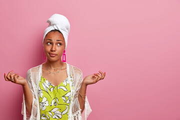 Wall Mural - Photo of hesitant ethnic woman spreads hands sideways, feels hesitant, cannot decide what to wear for date, wears wrapped towel on head, faces dilemma, poses against pink wall with blank empty space