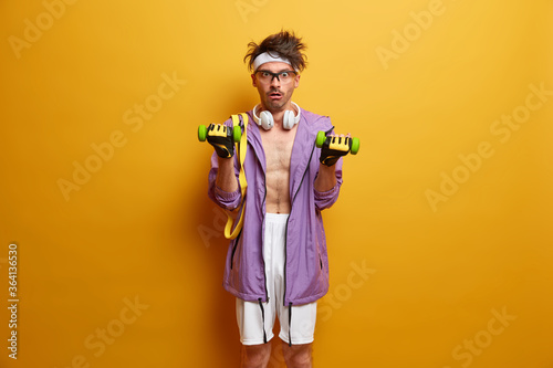 Cuadros en Lienzo Skinny man has aim of perfect muscular body, raises dumbbells and stares with sh