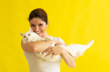 Wall Mural - Happy middle aged caucasian woman with a white cat on a yellow background. The female owner lovingly hugs her furry pet. Pets.