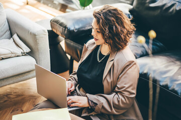 Caucasian brunette woman sitting near the sofa in living room and using laptop for freelance job.