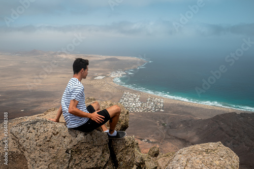 Cuadros en Lienzo Hombre joven contempla la playa de Famara desde la cima de una montaña
