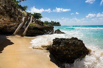 Waves crash into a large rock on the sandy shore and splash high in the air.