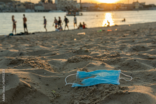 A used disposable mask (protect from COVID-19, Coronavirus) is on a beach, covered with sand, leading to bad consequences like pollution or contamination of the nature and water