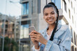 © ARUTA Images - Beautiful African American woman using cellphone, communication, walking on the street, looking at camera, smiling. Stylish hipster girl holding smartphone, listening to music outdoors
