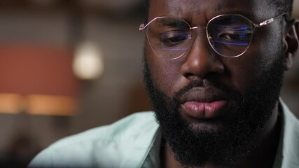Close-up of dark-skinned male programmer looking at screen of laptop, program code reflecting in his glasses. Serious african american software developer in eyeglasses working on computer