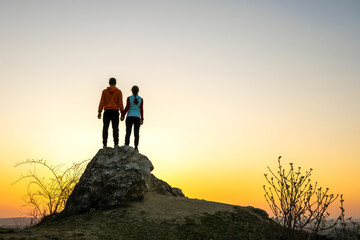 Canvas Print - Man and woman hikers standing on a big stone at sunset in mountains. Couple together on a high rock in evening nature. Tourism, traveling and healthy lifestyle concept.