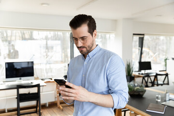 Wall Mural - Young Caucasian male employee in glasses look on cellphone screen text or message in office, concentrated businessman use modern smartphone gadget, browse wireless internet at workplace