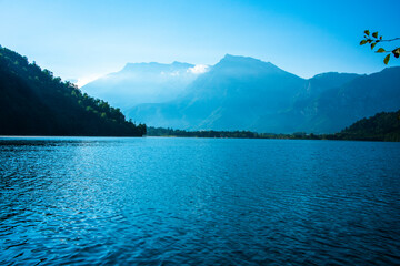 trees are reflected on Lake Levico with the mountains in the background in Trento, Italy