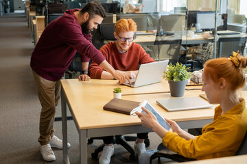 Leinwandbilder - Young bearded businessman pressing key of laptop keypad while helping colleague