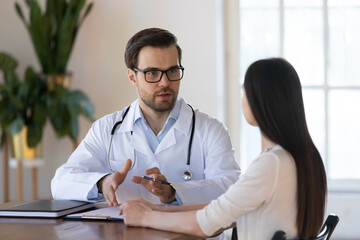 Young male doctor wearing white uniform with stethoscope consulting woman at meeting in clinic, gp discussing medical checkup results with female patient, giving treatment prescriptions, healthcare