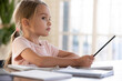 © fizkes - Close up thoughtful little girl sitting at desk, studying at home, pondering school homework, assignments, holding pencil, watching online video course, remote education, homeschooling