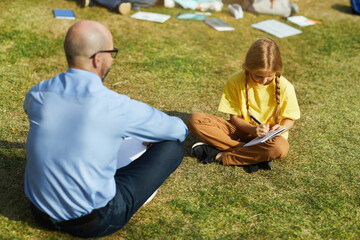 Wall Mural - full length portrait of blonde teenage girl sitting on green grass in sunlight and writing in notebo
