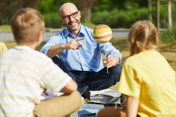 Wall Mural - portrait of bald male teacher pointing at planet model and smiling while talking to group of childre