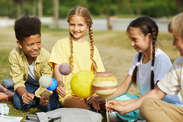 Wall Mural - multi-ethnic group of kids sitting on green grass and holding model planets while enjoying outdoor a