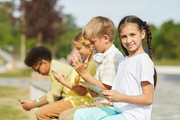 Wall Mural - Side view at multi-ethnic group of children using tablets and smartphones while sitting in row outdoors in sunlight, focus on smiling girl in foreground, copy space