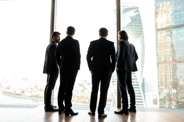 Full length successful young businessmen in formal suits standing near huge panoramic window, discussing working issues or partnership details in informal conversation enjoying cityscape in office.
