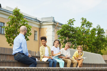 Wall Mural - multi-ethnic group of children listening to male teacher while sitting on bench and enjoying outdoor