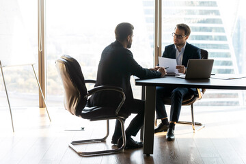 Wall Mural - Full length focused male hr manager in glasses and formal wear holding paper resume, involved in job interview with skilled male candidate applicant, sitting together at table in modern office.