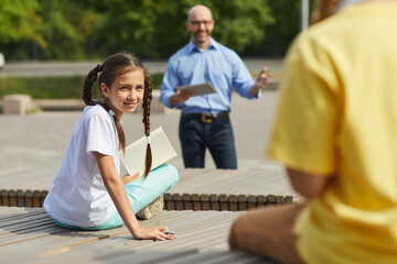 Wall Mural - Portrait of smiling teenage girl looking over at classmate while enjoying outdoor lesson in sunlight with male teacher in background, copy space
