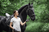 Fototapeta Konie - portrait of beautiful young woman standing near black friesian stallion in forest