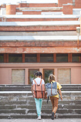 Wall Mural - Vertical back view portrait of two sisters going to school with backpacks while walking up stairs to big building, copy space