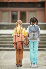 Wall Mural - Full length back view portrait of two cute schoolgirls with backpacks standing outdoors against school building and holding hands