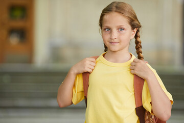 Wall Mural - Waist up portrait of cute freckled schoolgirl carrying backpack and looking at camera while posing outdoors against school building, copy space