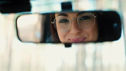 Wall Mural - Young woman sit in car during travelling break. Girl applying red or nude color lipstick on lips and look into fron view mirror. Smiling to her reflection and looking on camera.