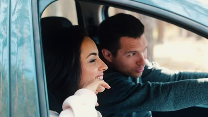 Wall Mural - Young man and woman riding together in car in forest. Slow motion of happy positive couple driving and enjoying travel. Guy pointing forward on beautiful nature outside car.